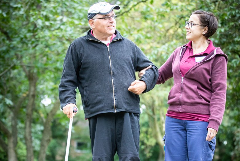 Ein älterer Mann mit Brille, schwarzer Jacke und Gehstock spaziert mit einer jüngeren Frau durch einen grünen Park. Sie trägt eine weinrote Kapuzenjacke und blaue Jeans und hält seinen Arm unterstützend. Beide schauen sich an und wirken freundlich und entspannt.