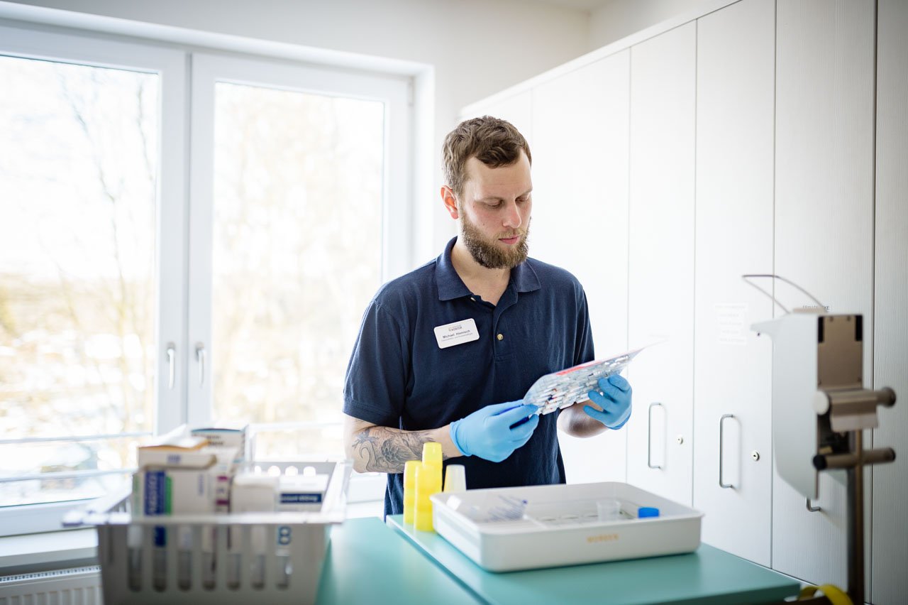 Ein Mann mit kurzem Bart und dunklem Poloshirt arbeitet konzentriert in einem hellen Raum. Er trägt blaue Einweghandschuhe und hält eine Spritze in der Hand, während er eine medizinische Substanz vorbereitet.