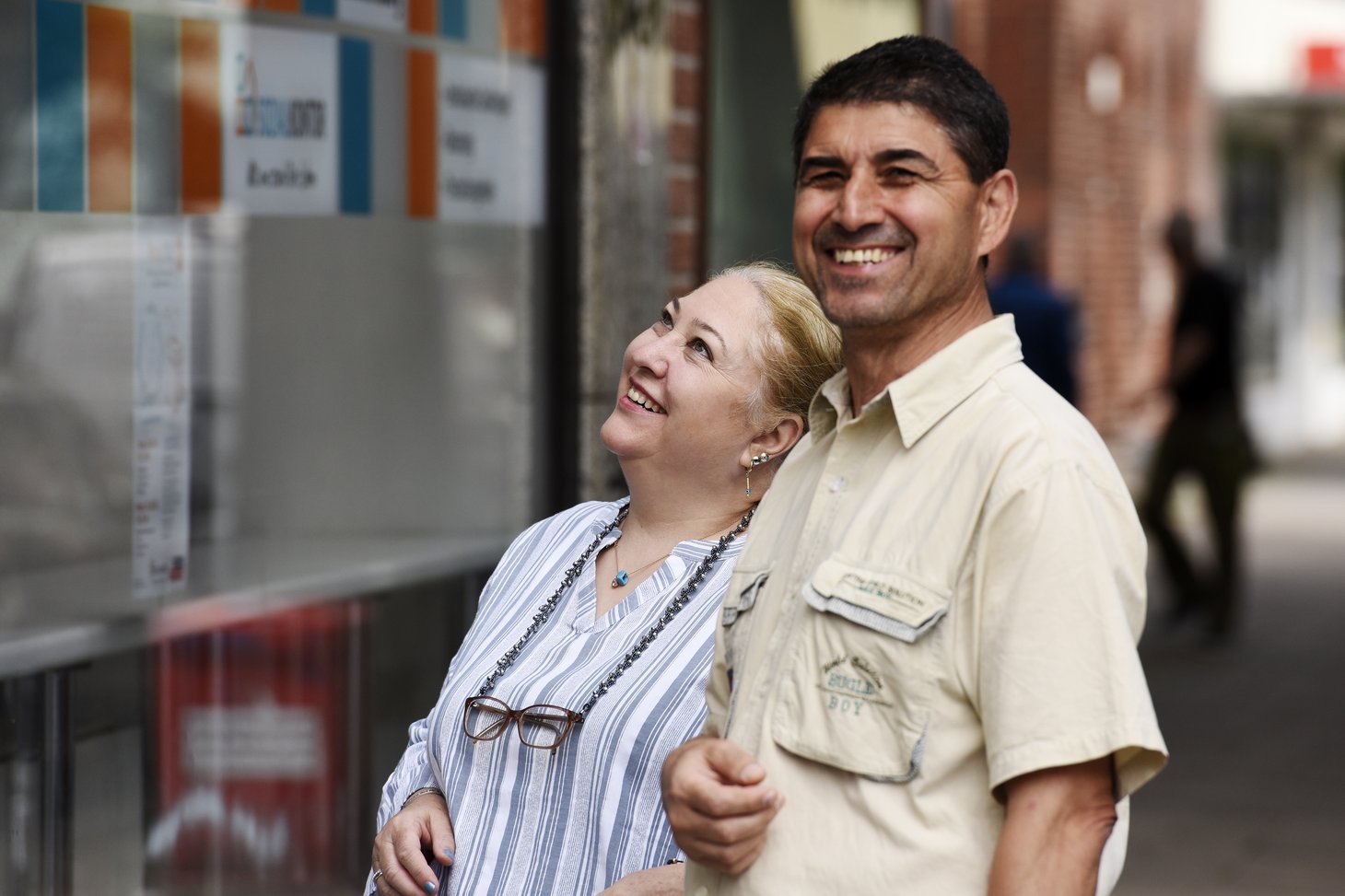 Das Bild zeigt zwei lachende Personen, eine Frau und einen Mann, die eng beieinander stehen. Die Frau lehnt sich zurück und lacht, während der Mann lächelnd in die Kamera blickt. Sie befinden sich in einer städtischen Umgebung mit Schaufenstern und Gebäuden im Hintergrund.