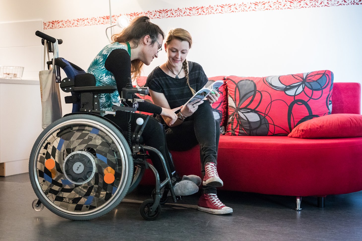 Eine Frau im Rollstuhl und eine weitere Frau sitzen in einem hellen Raum und schauen gemeinsam auf ein Tablet. 